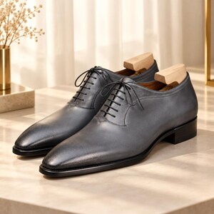 May include: A pair of grey leather oxford shoes with black laces, a pointed toe, and a low black heel. Each shoe features a smooth finish and is equipped with a light-colored wooden shoe tree inserted inside to maintain the shape of the footwear.