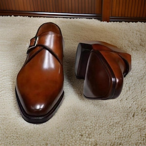 Könnte beinhalten: Ein Paar braune Monk-Strap-Schuhe aus Leder mit polierter Oberfläche. Die Schuhe haben einen Schnallenverschluss und eine spitze Zehenpartie.