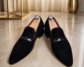 May include: A pair of black suede loafers featuring a pointed toe and a glossy black patent leather strap across the vamp. Each shoe includes a wooden shoe tree inserted inside, with a low stacked heel and a smooth, dark finish on the sole.