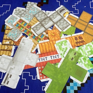 Puede incluir: Un conjunto de modelos de papercraft Minecraft imprimibles. Los modelos incluyen un bloque de TNT, un creeper, una estantería, un cofre y un bloque de diamante. Los modelos están impresos en papel blanco con diseños coloridos.