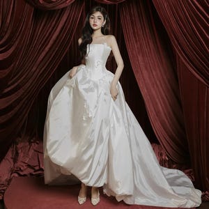 May include: A strapless white satin wedding gown with a full skirt and a long train. The dress features delicate floral embellishments on the bodice. The background includes burgundy velvet curtains and a red circular platform.