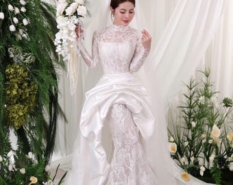 May include: A white lace wedding dress with long sleeves and a high neckline. The dress features a satin skirt with a draped design and a long train. The bride is holding a bouquet of white flowers.