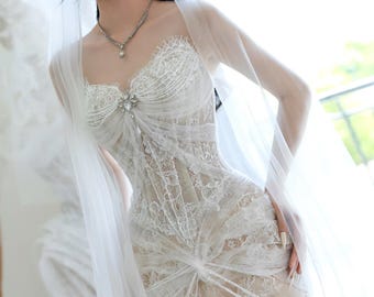 May include: A white lace wedding dress with a sweetheart neckline and a fitted bodice. The dress features intricate lace detailing and a flowing skirt. A sheer veil drapes over the shoulders. A silver necklace with a pendant and a jeweled brooch adorn the dress.