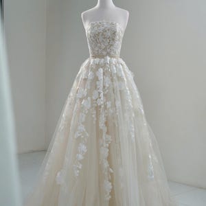 May include: An ivory strapless wedding dress on a mannequin. The bodice has detailed lace, and the full skirt is decorated with cascading floral appliqués. A gold crown sits on the mannequin's head.