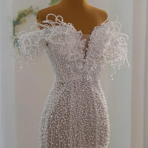 Op de afbeelding: Een witte, met kralen bezette trouwjurk op een mannequin. De jurk heeft een diepe V-hals en off-the-shoulder mouwen, beide versierd met ingewikkelde pareldetails. Het lijfje en de rok zijn bedekt met kleine, gelijkmatig verdeelde parels. Een gouden kroon staat op het hoofd van de mannequin.