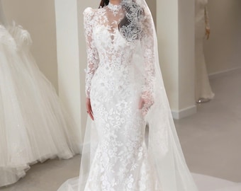 May include: A white, long-sleeved wedding dress with intricate floral lace detailing. The dress features a high neckline, a fitted bodice, and a mermaid silhouette. A sheer veil with lace trim completes the bridal look. The text "NH5I.TT006" is visible.