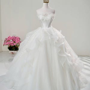 May include: A white wedding dress with a layered tulle skirt and a fitted bodice, displayed on a mannequin. The dress is topped with a gold crown. A bouquet of pink flowers is to the left. The dress is a classic bridal style.