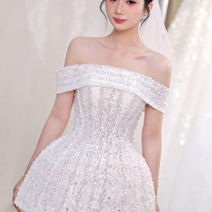May include: A white, off-the-shoulder bridal dress with intricate beadwork and a short skirt. The bodice and neckline are heavily embellished with sparkling beads, creating a glamorous look. The dress is designed for a wedding or special occasion.