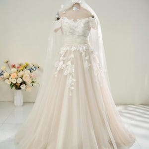 May include: A bridal gown with an off-the-shoulder neckline and a flowing tulle skirt. The ivory dress features floral appliqués and is paired with a matching veil. The gown is displayed on a mannequin.