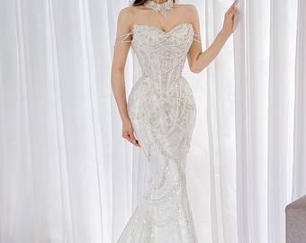 May include: A floor-length white strapless gown featuring a sweetheart neckline, intricate silver beadwork, and cascading crystal embellishments. The dress includes delicate off-the-shoulder straps and a fitted bodice that flares into a mermaid silhouette.