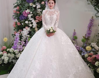 May include: A white lace wedding dress with long sleeves and a high neckline. The dress has a full skirt and is adorned with floral embroidery. The bride is holding a small bouquet of white flowers. The background features a floral arch with various colored flowers.