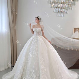 May include: A strapless white wedding gown with floral embellishments. The gown features a full skirt and a long train. A sheer veil and a jeweled headpiece complete the bridal look. A chandelier hangs in the background.