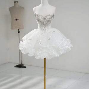 Könnte beinhalten: Ein weißes trägerloses Kleid mit herzförmigem Ausschnitt, verziert mit silbernen Perlen. Das Oberteil ist figurbetont, und der Rock hat ein geschichtetes, gerüschtes Design. Eine goldene Krone sitzt auf dem Kopf der Schaufensterpuppe. Das Kleid wird auf einem goldenen Ständer präsentiert.