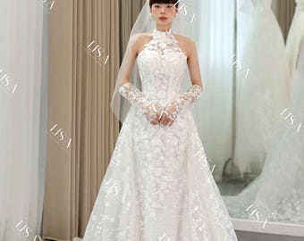 May include: A white wedding dress with a high neckline and floral lace detailing. The dress features a fitted bodice, a flowing skirt, and long lace gloves. A sheer veil is also present. The dress is displayed in a bridal shop.