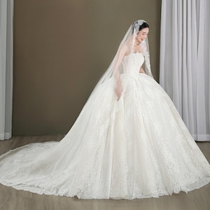May include: A strapless white wedding gown with a full skirt and a long train. The dress is adorned with intricate lace detailing. A sheer veil is draped over the head and shoulders.