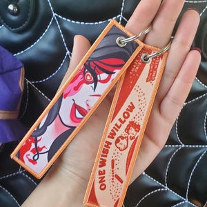 May include: Two rectangular fabric keychains with orange stitched borders and metal rings. One features a stylised illustration of a person with dark hair and red accents. The other is orange and white with the text ONE WISH WILLOW and small graphics.