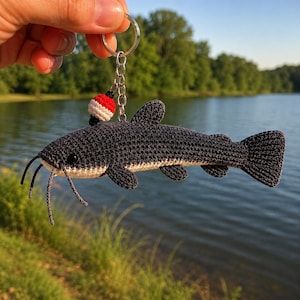 Puede incluir: Un llavero de bagre tejido a ganchillo con cuerpo gris oscuro, vientre color crema, bigotes largos y un pequeño flotador de pesca rojo y blanco unido a una cadena de anillo de metal plateado, mostrando puntadas detalladas.
