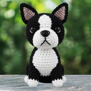 May include: A handmade crochet Boston Terrier dog figurine. The dog is primarily black and white, with a white chest, face, and paws. The ears are black with a tan interior. The dog is sitting upright, with a focused expression, and is made of soft yarn.