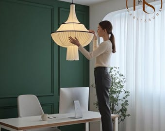 May include: A cream-colored fabric pendant light with a tiered, bell-shaped design and black trim accents. The fixture features a pleated texture and hangs from a thin black cord, suspended from the ceiling above a white desk and chair.