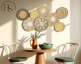 May include: A collection of woven wall decor in various circular designs, mounted on a light-colored wall. The decor is made of natural fibers in shades of beige and brown. A round wooden table with a terracotta pitcher, a teal bowl, and two chairs complete the scene.