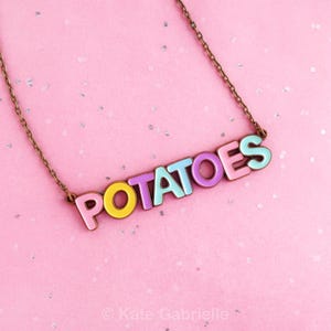 May include: A necklace featuring a metal chain and a pendant that spells out the word POTATOES in individual, colorful enamel letters. The letters are colored in shades of pink, yellow, purple, and light blue, attached to a thin bronze-toned chain.