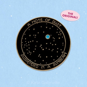 Pode incluir: Um broche redondo de esmalte preto com uma borda dourada e letras douradas que dizem A MOTE OF DUST SUSPENDED IN A SUNBEAM. O centro exibe um campo de pequenos pontos dourados representando estrelas e um único ponto circular azul brilhante.