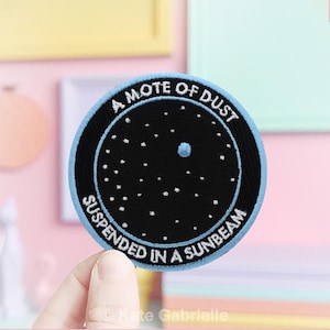 May include: Round embroidered patch with a black background and white star-like dots. The patch has a light blue border and white text that reads "A MOTE OF DUST SUSPENDED IN A SUNBEAM". The patch is circular.