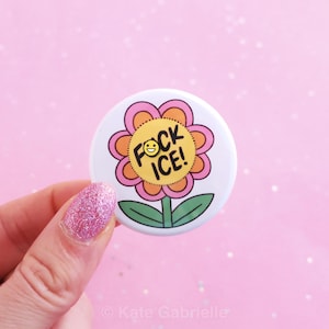 May include: A white button with a flower design. The flower has pink and orange petals, green leaves, and the text "FOCK ICE!" with a smiley face. The button is held by a hand with pink glitter nail polish.