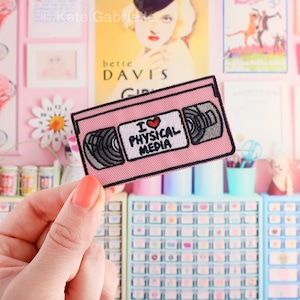 May include: A rectangular embroidered patch featuring a pink VHS cassette tape design. The center white label displays the black text I LOVE PHYSICAL MEDIA with a small red heart symbol. The tape reels are depicted in shades of grey and black.