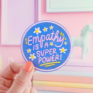 May include: A circular embroidered patch with a blue background and a light pink border. The text reads "Empathy IS A SUPER POWER!" in pink, surrounded by small white and yellow flowers and yellow stars, with a yellow line underneath the text.