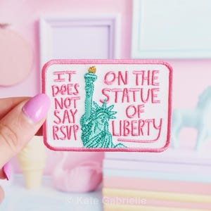 Puede incluir: Un parche rectangular con borde rosa y el texto "IT DOES NOT SAY RSVP ON THE STATUE OF LIBERTY". El parche presenta una imagen bordada de la Estatua de la Libertad en turquesa con una antorcha dorada.