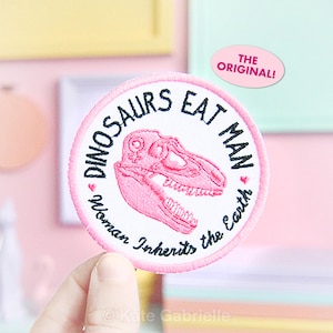 May include: A round, pink and white embroidered patch with the text "DINOSAURS EAT MAN" and "Woman Inherits the Earth". The patch features a pink dinosaur skull graphic. A pink oval sticker reads "THE ORIGINAL!"