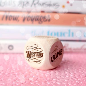 May include: A wooden die with the words "Western" and "Crime" etched on its sides. The die is light-colored and sits on a pink, glittery surface. Books are blurred in the background.