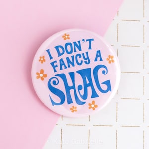 May include: A round, light pink button with the text "I DON'T FANCY A SHAG" in blue, with orange flower accents. The button is set against a pink and white background.