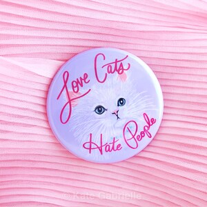May include: A round, light purple button with a white cat illustration and the text "Love Cats Hate People" in pink script. The button is set against a pink, pleated fabric background.