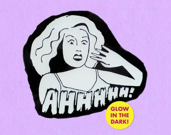 Op de afbeelding: Zwart-wit afbeelding van een vrouw met een geschokte uitdrukking, hand naar haar gezicht, en het woord "AHHHHH!" eronder. Een gele cirkel met de tekst "GLOW IN THE DARK!" staat rechtsonder.