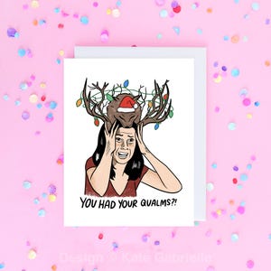 May include: A white greeting card featuring a cartoon illustration of a person with dark hair holding their head in distress. A brown creature wearing a Santa hat and large antlers wrapped in colorful Christmas lights sits on their head. Text reads: YOU HAD YOUR QUALMS?!