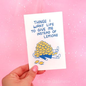 Puede incluir: Una tarjeta de felicitación blanca con texto azul que dice: "THINGS I WANT LIFE TO GIVE ME INSTEAD OF LEMONS." Debajo del texto hay una ilustración con contorno azul de una figura acostada, enterrada bajo un montón de limones amarillos.