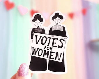 Pode incluir: Um autocolante a preto e branco com duas mulheres a segurar uma placa com a frase "VOTES FOR WOMEN". As mulheres usam vestidos longos pretos. O autocolante tem uma borda branca e é segurado por uma mão com verniz de unhas rosa brilhante.