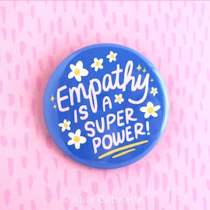 May include: A round, blue button with the words "Empathy is a Super Power!" in white script. The button is decorated with white flowers and yellow accents. The background is pink with a repeating pattern.