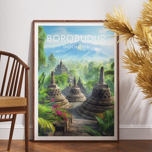 Può includere: Una stampa incorniciata raffigurante il tempio di Borobudur in Indonesia. L'opera d'arte mostra l'antico complesso del tempio buddista con una vegetazione lussureggiante, un cielo blu e il testo "BOROBUDUR INDONESIA". La stampa è esposta su un pavimento in legno.