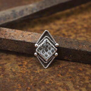 May include: A silver ring with a diamond-shaped design. The center of the ring has a textured pattern, and the edges are adorned with a rope-like texture and small silver beads. The ring is set against a rusty metal background.