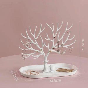 May include: White jewelry organizer in the shape of a tree with a deer base. The tree-shaped organizer is 21.5 cm tall and 24.5 cm wide, with an oval base that is 12.5 cm wide. Several pieces of jewelry are displayed on the branches.
