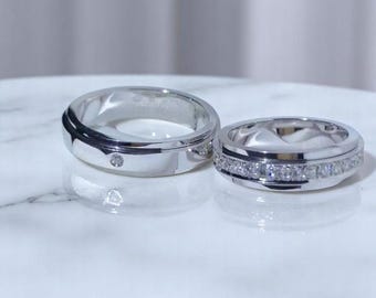 May include: Two silver rings. One ring has a single small diamond, the other has a row of small diamonds. Both rings are on a white surface. The rings are likely wedding or engagement rings.
