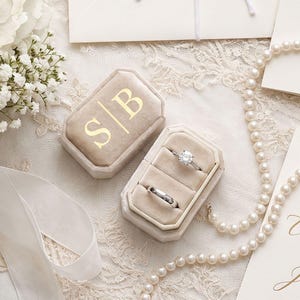 Pode incluir: Uma caixa de veludo bege para anéis com as letras douradas S|B, contendo dois anéis prateados, um com uma pedra central. A caixa está rodeada por um bouquet branco, um colar de pérolas, um envelope branco e artigos de papelaria.