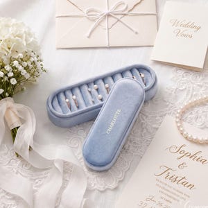 May include: A light blue velvet ring box with the name Charlotte embossed in gold, containing several rings. The box is surrounded by a white floral bouquet, a wedding invitation for Sophia and Tristan, a vow booklet, and a string of pearls.