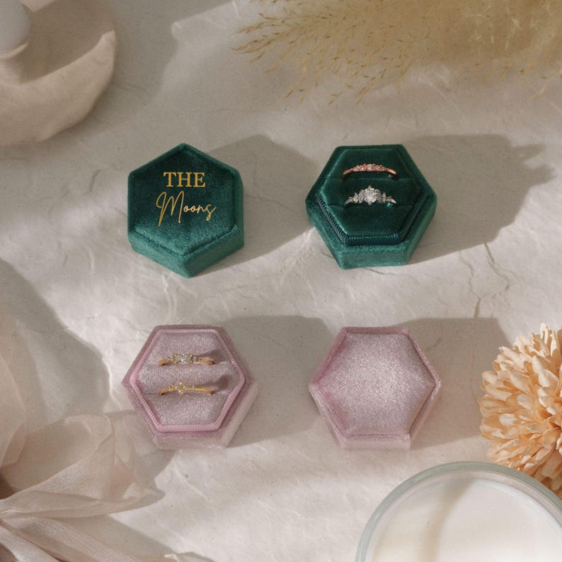 Puede incluir: Cuatro joyeros hexagonales de terciopelo en tonos verdes y rosas. Dos joyeros est&aacute;n abiertos, mostrando anillos con diamantes y piedras preciosas. Un joyero verde tiene el texto dorado "The Moons". Los joyeros est&aacute;n colocados sobre una superficie clara.