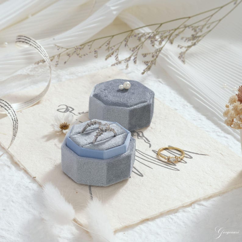 Puede incluir: Dos joyeros de terciopelo gris, uno abierto con un anillo dentro, y un anillo dorado con tres piedras. Los joyeros est&aacute;n sobre una superficie color crema con elementos decorativos. El joyero abierto tiene el interior azul claro.