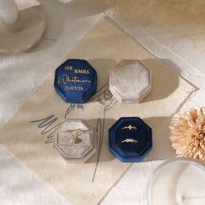 Puede incluir: Una variedad de joyeros octogonales de terciopelo azul y beige. Una caja azul tiene texto dorado que dice "MR. & MRS. Whitmore 20/07/25". Dos cajas azules contienen anillos.