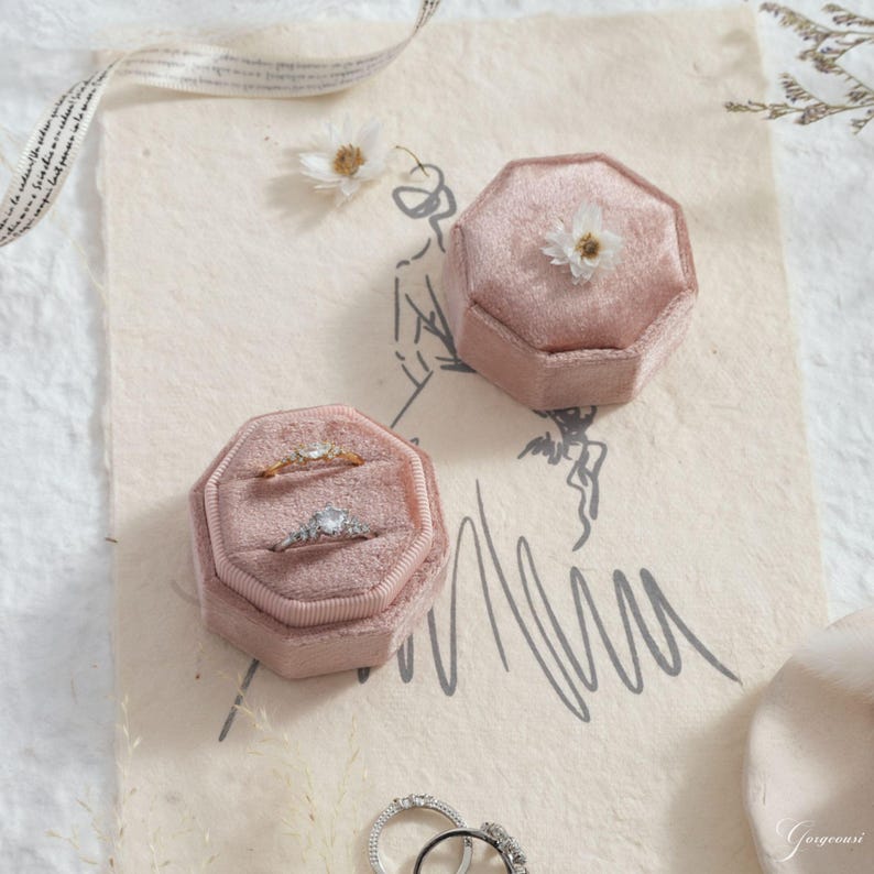 Puede incluir: Dos joyeros octogonales de terciopelo rosa, uno abierto con dos anillos, el otro cerrado con una peque&ntilde;a flor. Las cajas est&aacute;n sobre un papel color crema con un boceto y una cinta.
